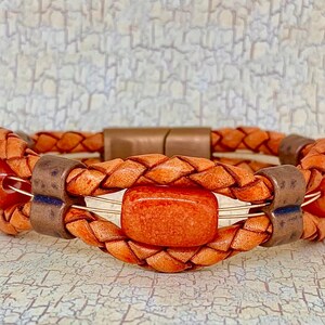 May include: Braided leather bracelet in shades of orange and brown. The bracelet features a central rectangular orange stone and is accented with copper-colored metal details. The design incorporates a woven leather pattern.