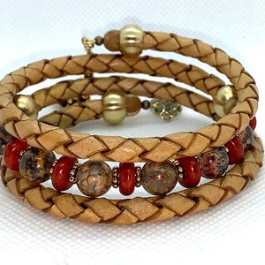 May include: A triple wrap leather bracelet with brown and red beads. The leather is a light tan color and the beads are a mix of brown, red, and gold.