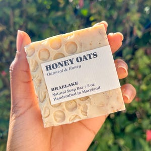 Puede incluir: Una barra de jabón blanca con un patrón de panal y el texto "HONEY OATS Oatmeal & Honey" y "BRAELAKE Natural Soap Bar | 5 oz Handcrafted in Maryland".