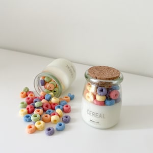 Puede incluir: Dos frascos de vidrio llenos de cereales de colores. Un frasco está en posición vertical con una tapa de corcho, el otro está volcado, derramando cereales. Los frascos tienen una etiqueta blanca con la palabra "CEREAL" impresa.