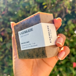 May include: A brown and green bar of soap with black specks. The soap is labeled "Energize Coffee Grounds" and "Braelake Natural Soap Bar | 5 oz Handcrafted in Maryland".