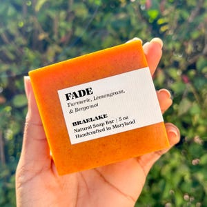 May include: A rectangular bar of orange soap with a white label that reads "FADE Turmeric, Lemongrass, & Bergamot BRAELAKE Natural Soap Bar | 5 oz Handcrafted in Maryland"