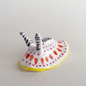 May include: A small ceramic sculpture of a sea slug with a white body, red and yellow spots, and black and white striped antennae.