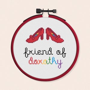 Puede incluir: Un aro de bordado blanco con costuras rojas alrededor del borde. El aro contiene un diseño de punto de cruz de un par de zapatillas rojas rubí con un diseño de arcoíris. El texto "friend of dorothy" está cosido en colores arcoíris debajo de las zapatillas.
