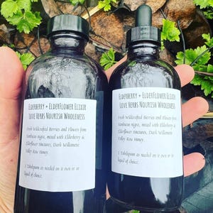 May include: Two dark glass bottles with dropper tops, labeled "Elderberry + Elderflower Elixir Love Herbs Nourish Wholeness". The labels describe the ingredients as fresh wildcrafted berries and flowers from Sambucus nigra, mixed with elderberry and elderflower tinctures, Dark Willamette Valley Raw Honey. The bottles are held in a hand.