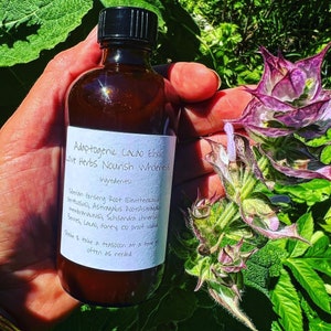 May include: A brown glass bottle with a white label that reads "Adaptogenic Cacao Elixir Love Herbs Nourish Wholeness" and lists the ingredients. The bottle is being held in a hand with a green plant in the background.