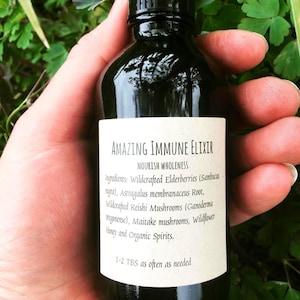 May include: A dark glass bottle with a white label that reads "AMAZING IMMUNE ELIXIR" and lists the ingredients: Wildcrafted Elderberries, Astragalus membranaceus Root, Wildcrafted Reishi Mushrooms, Maitake mushrooms, Wildflower Honey and Organic Spirits. The label also states "NOURISH WHOLENESS" and "1-2 TBS as often as needed".