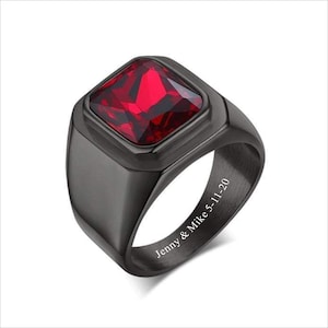 May include: A black ring with a large, square, red gemstone. The inside of the band is engraved with "Jenny & Mike 5-11-20".