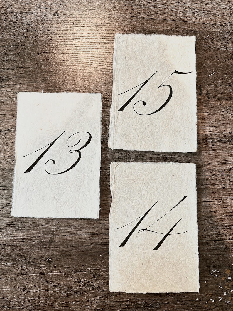 May include: Three white paper cards with black calligraphy numbers 13, 14, and 15. The cards are on a wood surface.