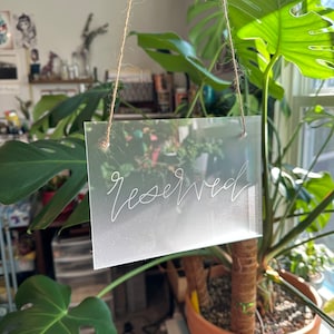 May include: A clear acrylic sign with the word "reserved" written in white lettering. The sign is hanging from a string.