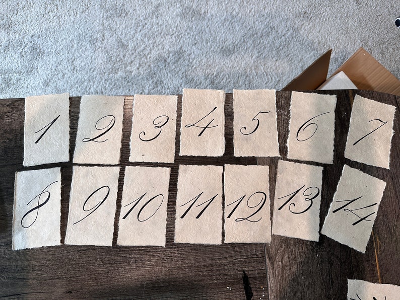May include: A set of 14 handmade paper cards with handwritten numbers from 1 to 14. The cards are beige and have a rough, textured surface.