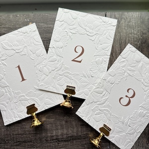 May include: Three white rectangular table number cards featuring embossed floral patterns and centered copper-colored numerals one, two, and three. Each card is held upright by a small, polished gold-toned metal stand at the bottom edge.