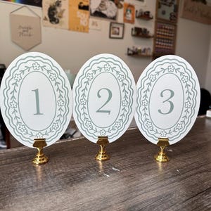 May include: Three white oval table number cards with a floral border and the numbers 1, 2, and 3 in a light gray font. Each card is held by a gold-colored metal stand. The cards are on a wooden surface.