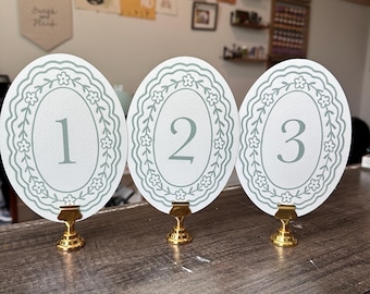 Números de mesa ovalados impresos en color salvia sobre papel grueso, letreros de boda con diseño floral de jardín.