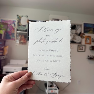 May include: A white paper sign with torn edges, featuring elegant calligraphy that reads "Please sign our photo guestbook." Additional text includes "Snap a photo. Place it in the book. Leave us a note." Signed "Love, Callie & Bryce."