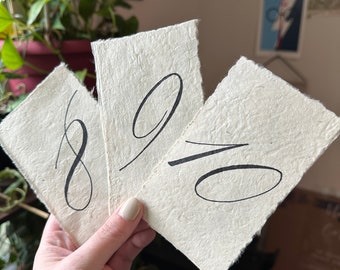 Números de mesa hechos a mano, con barbas y rasgados, sobre papel hecho a mano, números de mesa con caligrafía manuscrita sobre papel texturizado.