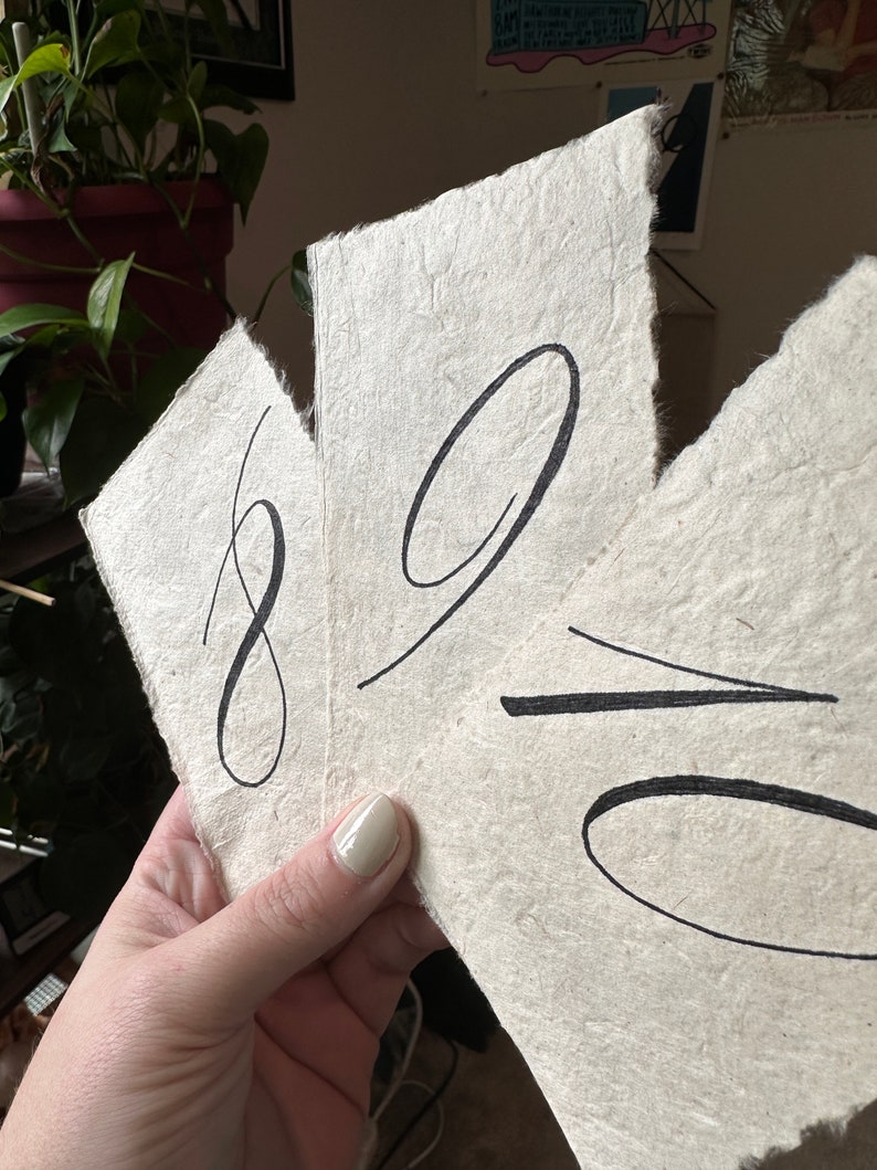 May include: Close-up of a hand holding two pieces of handmade paper with black ink calligraphy. The paper is textured and has a rough edge. The calligraphy says "89 = ".