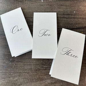 May include: Three rectangular white paper table cards with folded edges. Each card features black printed text in a cursive script font, displaying the words One, Two, and Three respectively, centered on the front face of each individual card.