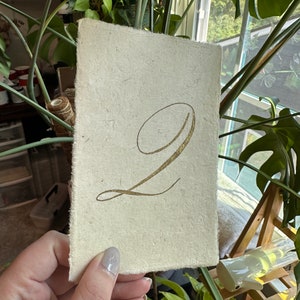 May include: A white card with a gold number "2" written in calligraphy. The card has a rough, textured surface.