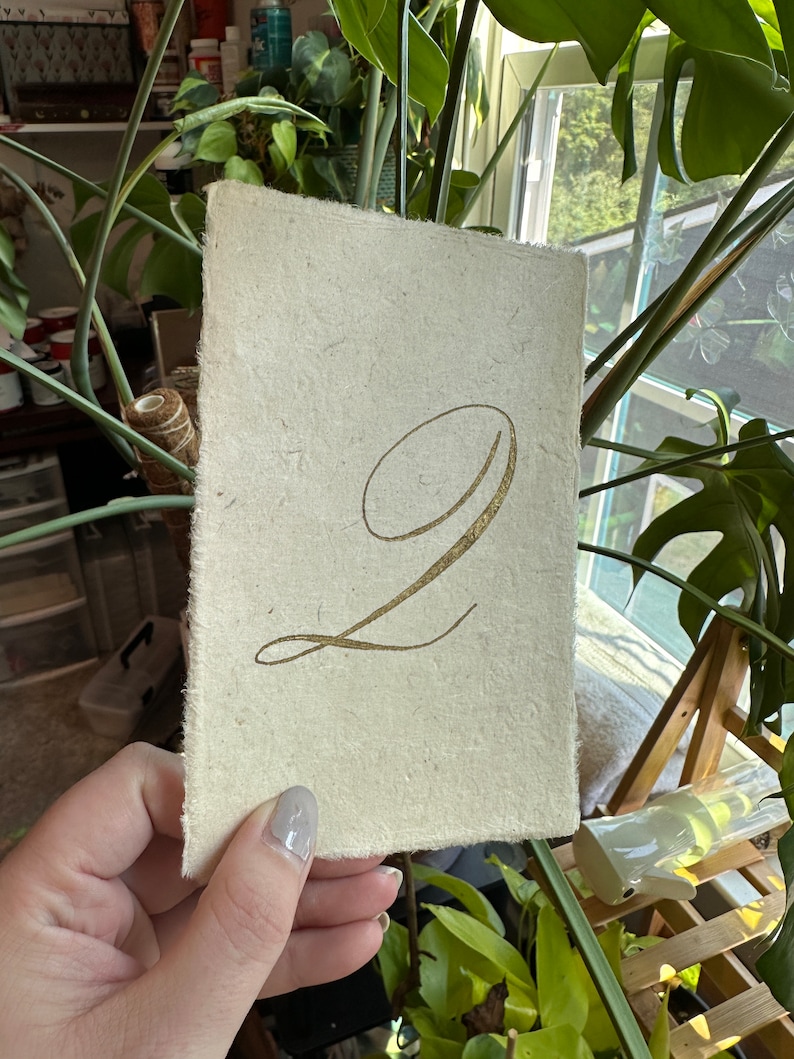 May include: A white card with a gold number "2" written in calligraphy. The card has a rough, textured surface.