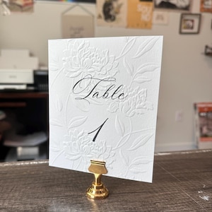 May include: A white rectangular table number card featuring an embossed floral pattern and the black printed text "Table 1" in a script and serif font. The card is held upright by a small, metallic gold-colored circular base stand.