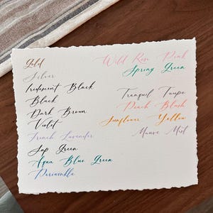 Puede incluir: Un papel blanco con muestras de caligraf&iacute;a manuscrita en varios colores, incluyendo oro, negro y violeta. El texto incluye nombres de colores como "Wild Rose Pink" y "Aqua Blue Green". El papel tiene un borde irregular y descansa sobre una superficie de madera.