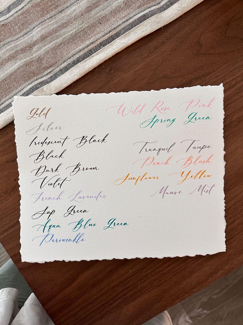 Puede incluir: Un papel blanco con muestras de caligraf&iacute;a manuscrita en varios colores, incluyendo oro, negro y violeta. El texto incluye nombres de colores como "Wild Rose Pink" y "Aqua Blue Green". El papel tiene un borde irregular y descansa sobre una superficie de madera.