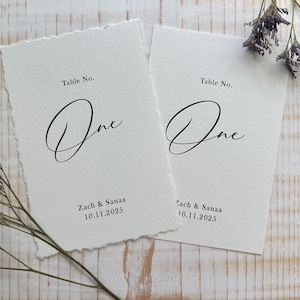 May include: Two off-white rectangular table number cards with deckled edges. The text reads Table No. One, Zach & Sanaa, and 10.11.2025 in black ink. The cards feature a script font for the word One and a serif font for the remaining text.