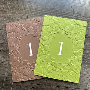 May include: Two rectangular table number cards, one in muted brown and one in bright lime green, both featuring an embossed floral border surrounding a smooth central area with the white printed number 1 centered on each card.