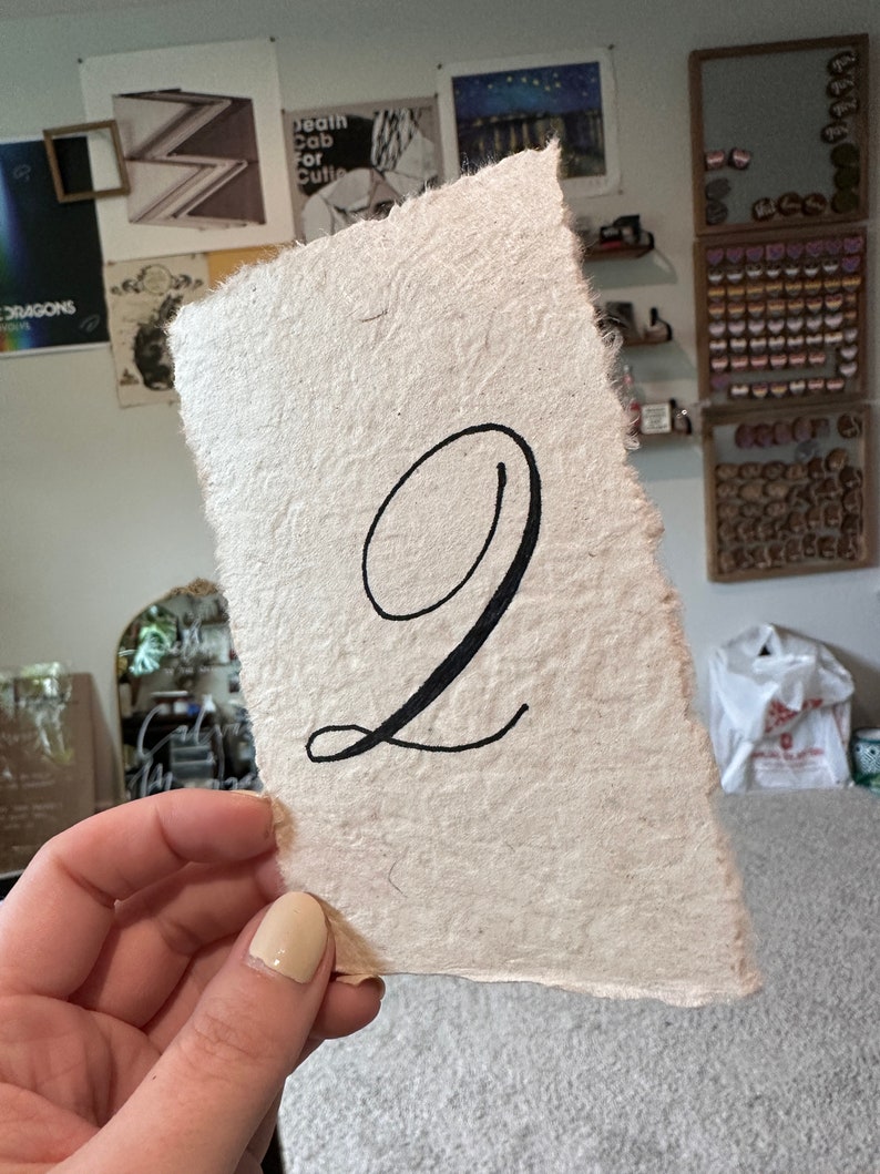 May include: A white, textured paper card with the number "2" written in black calligraphy.