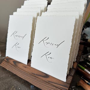 Puede incluir: Un expositor de madera que contiene varias tarjetas de papel blanco con bordes irregulares. Las tarjetas est&aacute;n impresas con las palabras "Reserved Row" en elegante caligraf&iacute;a negra. La madera tiene un acabado oscuro y te&ntilde;ido.