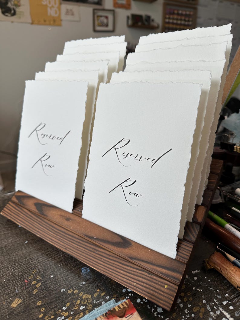 Puede incluir: Un expositor de madera que contiene varias tarjetas de papel blanco con bordes irregulares. Las tarjetas est&aacute;n impresas con las palabras "Reserved Row" en elegante caligraf&iacute;a negra. La madera tiene un acabado oscuro y te&ntilde;ido.