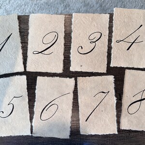 May include: Eight handmade paper cards with handwritten numbers 1 through 8 in black ink. The cards have a rough, textured surface and uneven edges.