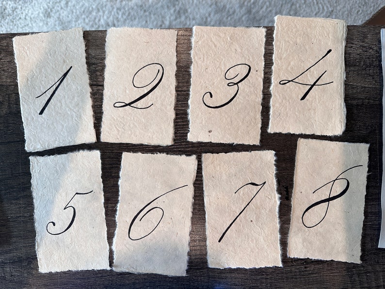 May include: Eight handmade paper cards with handwritten numbers 1 through 8 in black ink. The cards have a rough, textured surface and uneven edges.