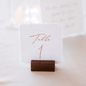 May include: A white card with gold lettering that reads "Table 1" is displayed on a wooden stand. The card is on a white tablecloth.