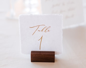 May include: A white card with gold lettering that reads "Table 1" is displayed on a wooden stand. The card is on a white tablecloth.