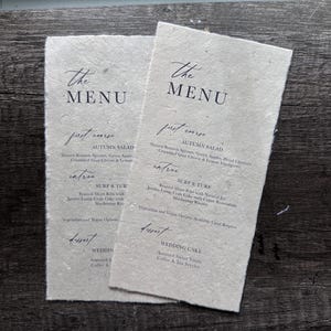 May include: Two beige paper menus with the text "The Menu" at the top. The first menu lists the following items: Autumn Salad, Surf & Turf, Wedding Cake. The second menu lists the following items: Autumn Salad, Surf & Turf, Wedding Cake.