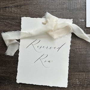 Puede incluir: Una tarjeta blanca con las palabras "Reserved Row" en caligraf&iacute;a elegante. La tarjeta tiene un borde irregular y est&aacute; atada con una cinta color crema. El fondo es una superficie de madera oscura.