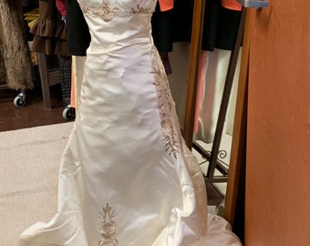 May include: A white satin wedding dress with gold embroidery. The dress has a sweetheart neckline and a fitted bodice. The skirt is full and flowing, with a train.