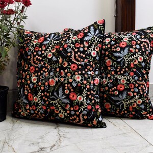 May include: Two black square throw pillows with floral embroidery in shades of pink, red, and blue. The pillows are on a white tiled floor.