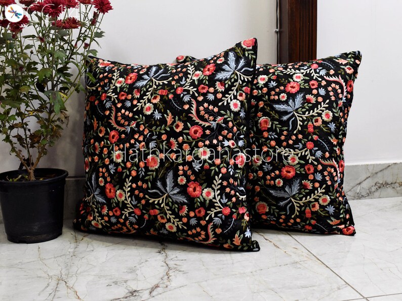 May include: Two black square throw pillows with floral embroidery in shades of pink, red, and blue. The pillows are on a white tiled floor.