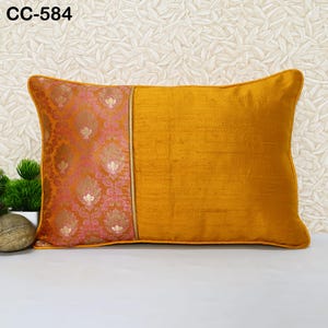 Può includere: Un cuscino rettangolare con un tessuto giallo senape su un lato e un tessuto con motivo floreale rosa e oro sull'altro lato. Il cuscino è su una superficie bianca con uno sfondo beige.