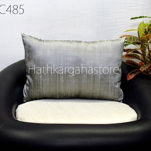 May include: A silver cushion with a subtle sheen sits on a black leather armchair. The cushion is placed on a white cushion. The armchair is in front of a white wall with a green plant in the background. The text "Hathkargahastore" is visible on the cushion.