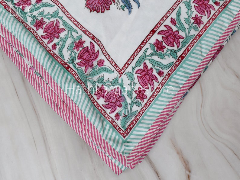 May include: Close-up of a white fabric with a pink and green floral print. The fabric is folded in layers, showing the pattern repeating. The fabric has a striped border in pink and green.