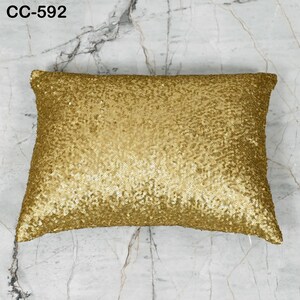 Puede incluir: Cojín rectangular dorado con lentejuelas. El cojín está cubierto de pequeñas y brillantes lentejuelas doradas. El texto "CC-592" está en la esquina superior izquierda. Está sobre una superficie de mármol.