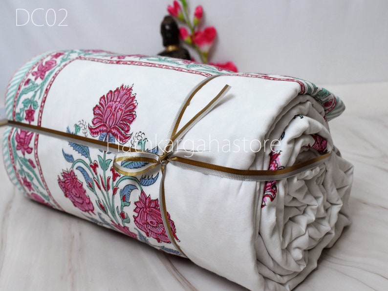 May include: A white cotton quilt with a floral print in shades of pink, green, and blue. The quilt is rolled up and tied with a gold ribbon. The text "DC02" is visible in the top left corner of the image.