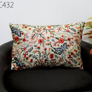 Puede incluir: Una almohada blanca con un diseño de bordado floral con flores rojas, rosas y azules sobre un fondo blanco. La almohada está sentada en un sofá negro.