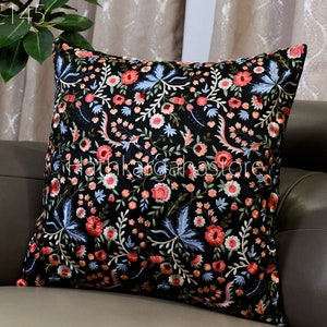 May include: Black velvet pillow with embroidered floral pattern in shades of pink, blue, and white. The pillow is on a brown leather couch.