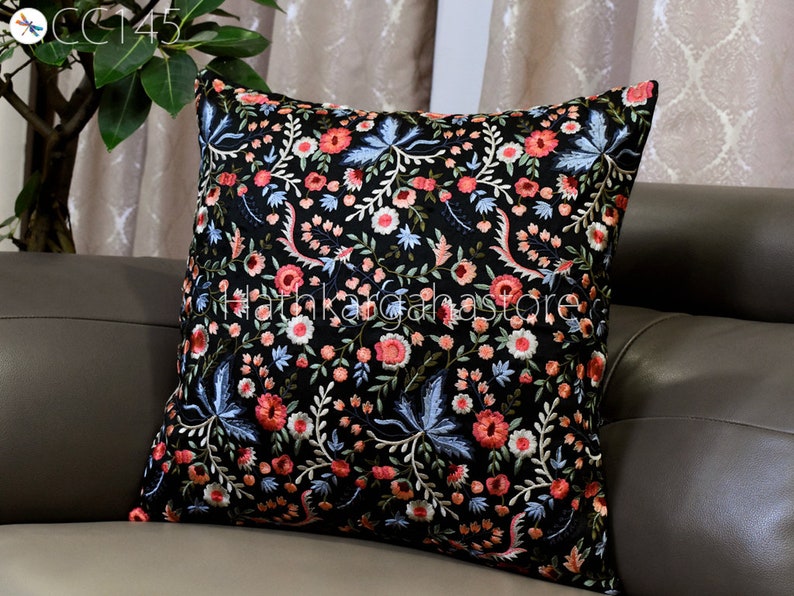 May include: Black velvet pillow with embroidered floral pattern in shades of pink, blue, and white. The pillow is on a brown leather couch.