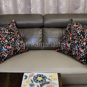 May include: Two black throw pillows with floral embroidery in shades of pink, blue, and white on a brown leather sofa. The pillows are arranged on the sofa with the floral patterns facing outward.